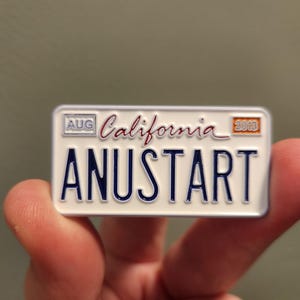 Puede incluir: Un pin rectangular que se asemeja a una placa de matr&iacute;cula de California. El pin es blanco con letras azules que dicen "ANUSTART". La palabra "California" est&aacute; escrita en cursiva. El pin tambi&eacute;n tiene el mes "AUG" y el a&ntilde;o "2013".