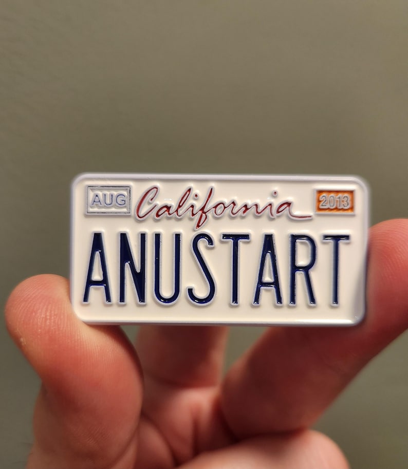 Puede incluir: Un pin rectangular que se asemeja a una placa de matr&iacute;cula de California. El pin es blanco con letras azules que dicen "ANUSTART". La palabra "California" est&aacute; escrita en cursiva. El pin tambi&eacute;n tiene el mes "AUG" y el a&ntilde;o "2013".