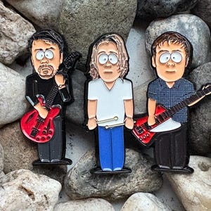 May include: Three cartoon figures with black outlines, each holding a musical instrument. The figure on the left is wearing a black shirt and holding a red electric guitar. The figure in the middle is wearing a white shirt and blue jeans and holding drumsticks. The figure on the right is wearing a blue and white plaid shirt and holding a red electric guitar.