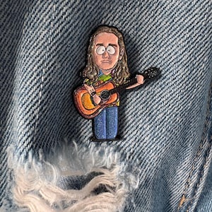 Può includere: Una spilla smaltata colorata raffigurante un musicista dei cartoni animati con i capelli lunghi che suona una chitarra acustica. La spilla ha un design dettagliato con colori vivaci ed è ambientata su una giacca di jeans.