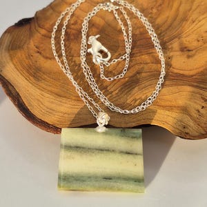 May include: A silver necklace featuring a square pendant in shades of light green and white, with horizontal stripes. The chain is fine, and the clasp is intricately designed. The pendant hangs against a wooden background.