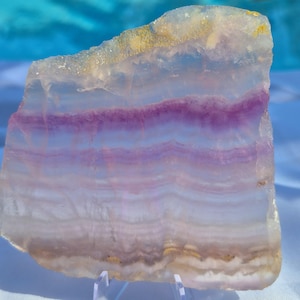 May include: A translucent, polished slab of fluorite mineral featuring horizontal bands of purple, lavender, and white. The stone has a jagged, natural top edge and a smooth, flat base, displaying internal crystalline structures and light refraction.