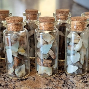 Könnte beinhalten: Eine Sammlung kleiner Glasflaschen, gefüllt mit verschiedenen bunten Steinen und Kristallen. Jede Flasche hat einen Korken. Die Steine sind in Blau-, Braun- und Cremetönen gehalten. Die Flaschen sind auf einer strukturierten Oberfläche angeordnet.