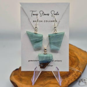 Op de afbeelding: Een sieradenset met een hangerketting en bijpassende oorbellen. De set toont blauwgroene edelsteenstukken met zilveren kettingen. De kaart erachter luidt "Tones Stones Sooke, British Columbia, gemstone, Silver". De set wordt op een houten standaard gepresenteerd.