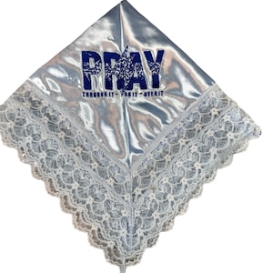 May include: A light blue satin head covering with white lace trim. The word "PRAY" is embroidered in dark blue, with the phrase "THROUGH IT - ON IT - OVER IT" below. The head covering is square-shaped.