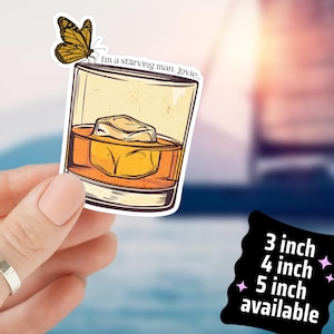 May include: A sticker featuring a whiskey glass with an ice cube and a butterfly. The text "I'm a starving man, Jovie" is on the glass. Available in 3, 4, and 5 inch sizes. The sticker is held in a hand.