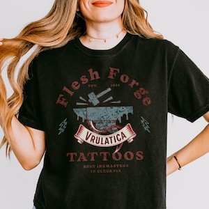 Puede incluir: Camiseta negra con un gráfico desgastado que presenta el texto "Flesh Forge Tattoos" y "Vrulatica". El diseño incluye un yunque, un martillo y una pancarta. La camiseta también tiene el texto "Best Inkmasters in Clecania".