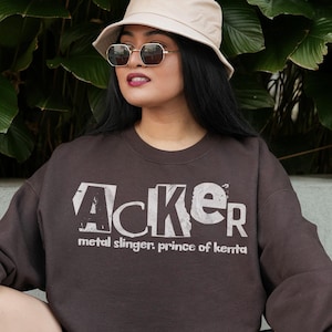 Peut inclure: Un sweat-shirt marron avec le mot "Acker" en blanc vieilli. Le texte "metal slinger, prince of kenta" est en dessous. Le sweat-shirt a des manches longues et un col rond.