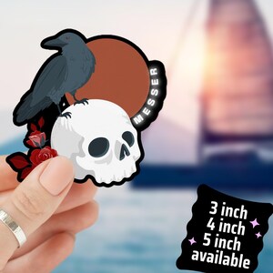 May include: A sticker featuring a raven perched on a white skull with red roses. The sticker has the word "MESSER" and is available in 3, 4, and 5-inch sizes. The sticker is held in a hand.