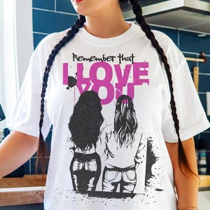 May include: White t-shirt with the text "Remember that I Love You" in black and pink. The graphic features two figures with long hair. The shirt is a casual, oversized style.