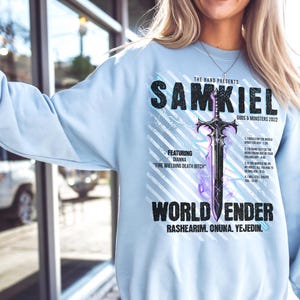May include: A light blue sweatshirt with a graphic print of a sword and the text "The Hand Presents Samkiel Gods & Monsters 2022 Featuring Dianna "Fire Welding Death Bitch" World Ender Rashearim. Onuna. Yejedin."