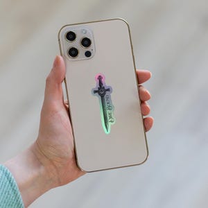 May include: A holographic sticker of a sword with the text "Dire Bound" on a smartphone. The sticker has a gradient of colors, including pink, green, and blue. The smartphone is gold-colored.