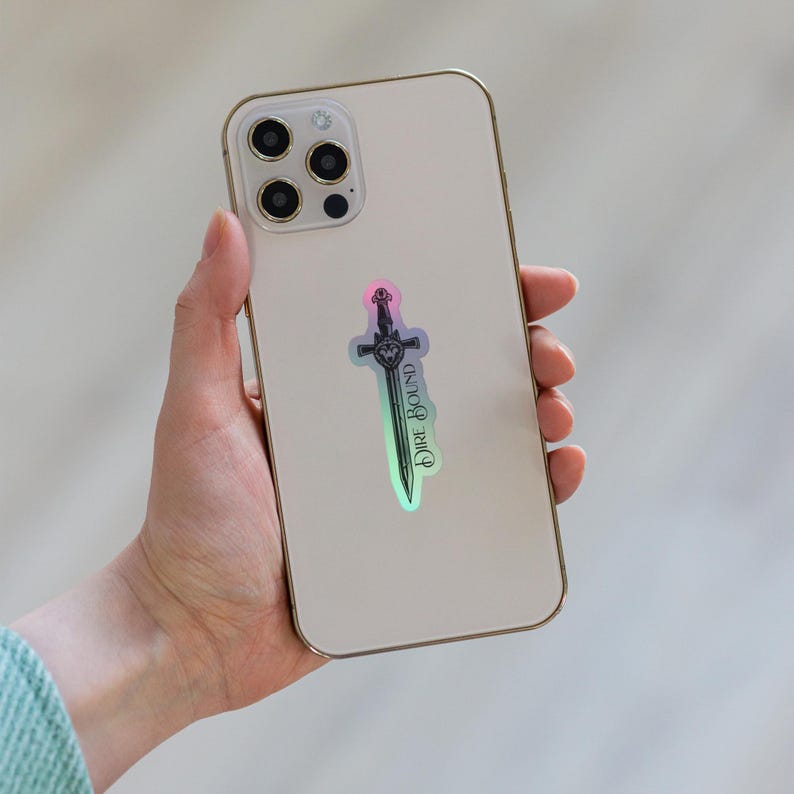 May include: A holographic sticker of a sword with the text "Dire Bound" on a smartphone. The sticker has a gradient of colors, including pink, green, and blue. The smartphone is gold-colored.