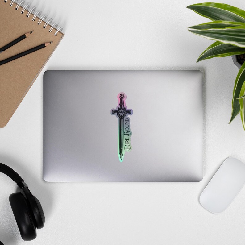 May include: A sticker featuring a sword design with a pink, green, and purple gradient. The text "Dire Bound" is vertically aligned on the blade. The sticker is on a laptop, with a notebook, pencils, headphones, a mouse, and a plant in the background.