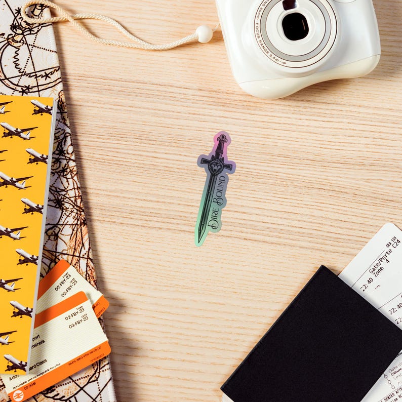 May include: A holographic sticker in the shape of a sword with the text "DIRE BOUND" printed vertically. The sticker has a gradient of colors, including purple, green, and pink. The sticker is on a light wood surface, with travel documents and a camera.