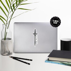 May include: A sticker featuring a detailed illustration of a sword with the words "Dire Bound" in a stylized font. The sticker is approximately 5 inches in size and is on a laptop.