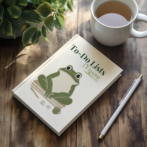 May include: A white notebook with the text "To-Do Lists I'll Ignore" and a green frog illustration. The notebook is on a wooden surface with a cup of tea and a pen.