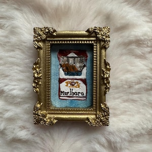 May include: A miniature painting of a Marlboro cigarette pack in a gold ornate frame. The painting features the iconic red and white packaging with the brand name. The frame has a detailed, vintage design, adding to the artwork's aesthetic.