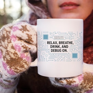 Pode incluir: Caneca de cerâmica branca com um design de placa de circuito e o texto "RELAX, BREATHE, DRINK, AND DEBUG ON." A caneca é segurada por uma pessoa que usa luvas estampadas em rosa e marrom.