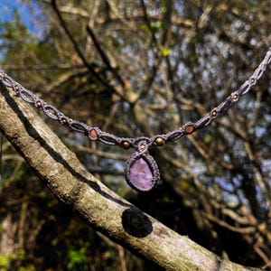 Peut inclure: Un collier avec un pendentif en pierre précieuse violette, un cordon en macramé et de petites perles dorées. Le collier est suspendu à une branche d'arbre.