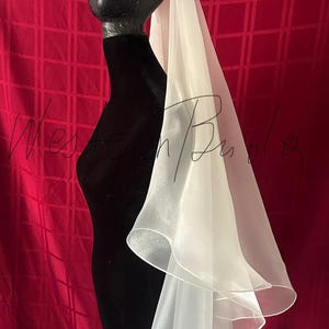 Op de afbeelding: Een witte, gelaagde sluier gedrapeerd over een zwarte mannequin hoofd en torso. De sluier heeft een zachte, vloeiende textuur en een delicate, gerimpelde rand. De achtergrond is een dieprode stof met een ruitpatroon.