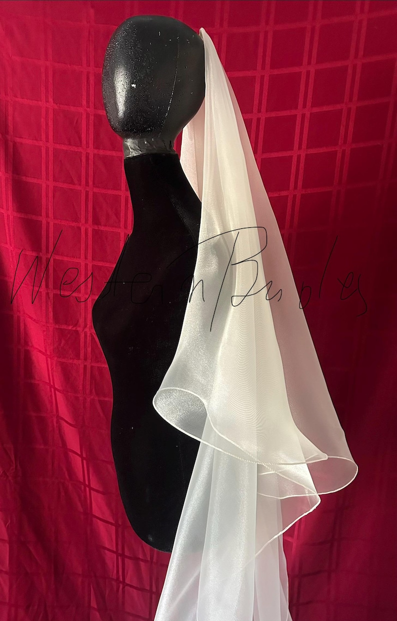 Op de afbeelding: Een witte, gelaagde sluier gedrapeerd over een zwarte mannequin hoofd en torso. De sluier heeft een zachte, vloeiende textuur en een delicate, gerimpelde rand. De achtergrond is een dieprode stof met een ruitpatroon.
