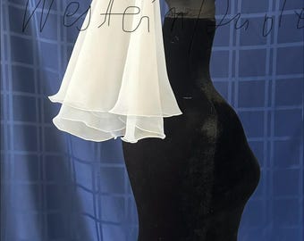 May include: A white, layered veil is draped over a black mannequin head and torso. The veil has a soft, flowing appearance with multiple tiers. The background is a blue fabric with a grid pattern. The text "Western Paola" is written on the image.