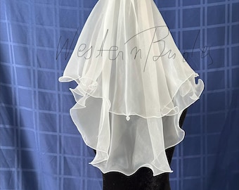 May include: A white, two-tiered bridal veil with a ruffled edge. The veil is made of a sheer fabric and is displayed on a black mannequin against a blue backdrop. The text "Western Bubbles" is written on the veil.