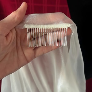 Op de afbeelding: Een doorschijnende witte sluier met een kam bevestigd. De sluier is gemaakt van een lichte, vloeiende stof. De kam heeft zilverkleurige tanden. De achtergrond is een mix van rood en zwart.