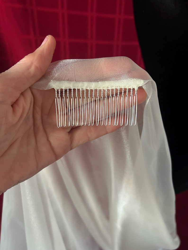 Op de afbeelding: Een doorschijnende witte sluier met een kam bevestigd. De sluier is gemaakt van een lichte, vloeiende stof. De kam heeft zilverkleurige tanden. De achtergrond is een mix van rood en zwart.