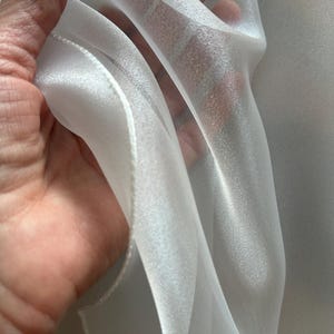 Op de afbeelding: Close-up van transparante witte stof, mogelijk organza of voile. De stof wordt omhoog gehouden en toont zijn delicate textuur en doorschijnendheid. Het materiaal heeft een subtiele glans, wat duidt op een hoogwaardige textiel, ideaal voor gordijnen of kleding.