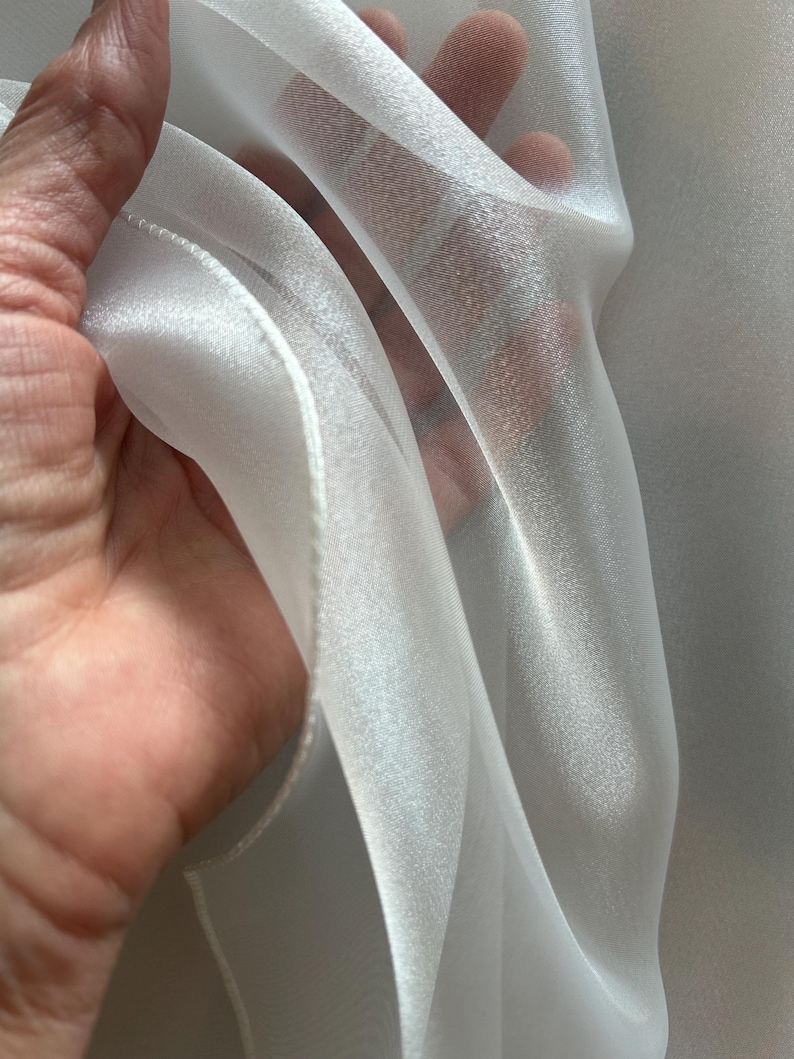 Op de afbeelding: Close-up van transparante witte stof, mogelijk organza of voile. De stof wordt omhoog gehouden en toont zijn delicate textuur en doorschijnendheid. Het materiaal heeft een subtiele glans, wat duidt op een hoogwaardige textiel, ideaal voor gordijnen of kleding.