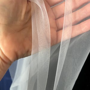 Puede incluir: Primer plano de tela de tul blanca transparente. La tela se sostiene, revelando su delicado tejido abierto. El tul tiene una apariencia suave y fluida, con un ligero brillo. La tela se utiliza probablemente para ropa de novia, velos o fines decorativos.