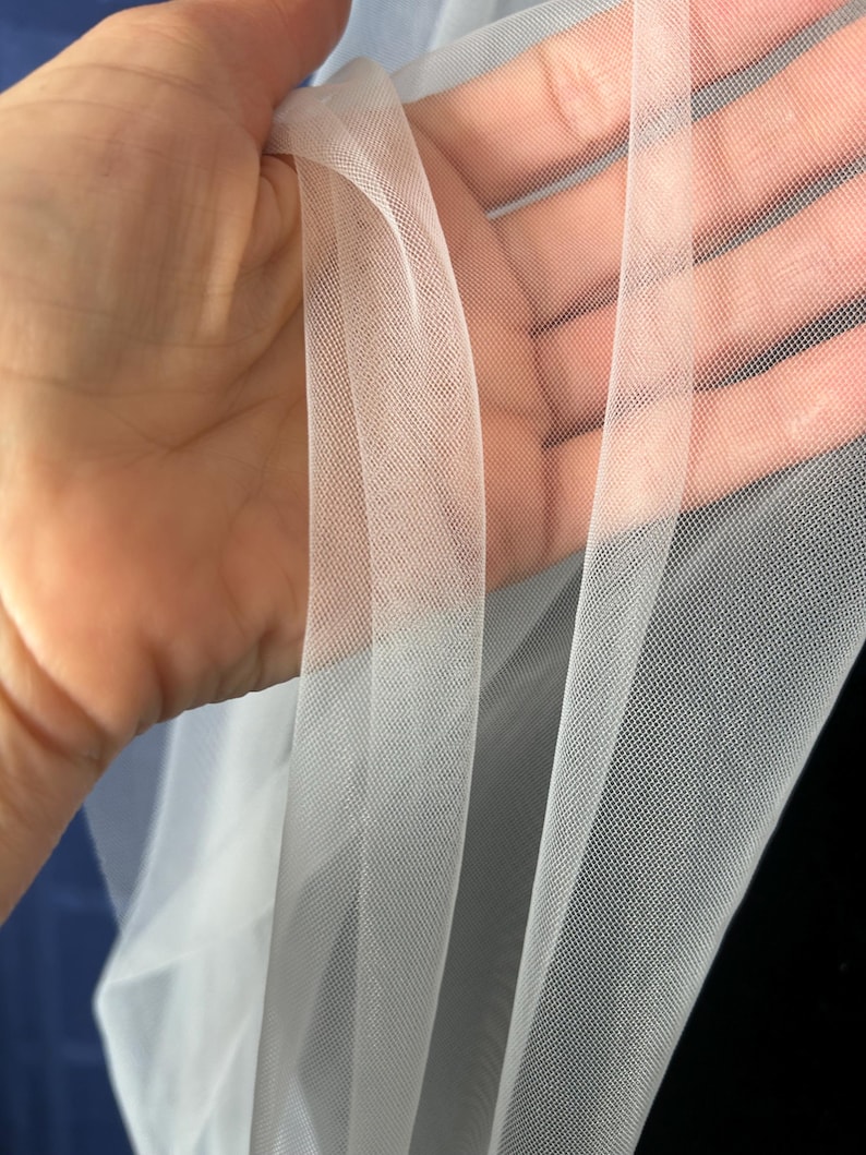 Puede incluir: Primer plano de tela de tul blanca transparente. La tela se sostiene, revelando su delicado tejido abierto. El tul tiene una apariencia suave y fluida, con un ligero brillo. La tela se utiliza probablemente para ropa de novia, velos o fines decorativos.