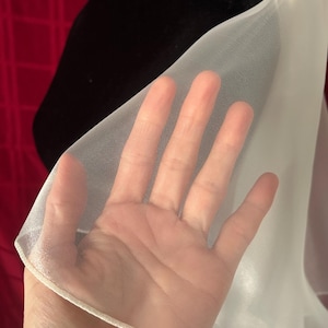 Op de afbeelding: Een close-up van een doorschijnende witte stof, mogelijk een sjaal of omslagdoek, tegen een zwarte achtergrond. De stof is dun en doorschijnend, met een hand erachter zichtbaar. De randen zijn afgewerkt met een delicate witte bies.