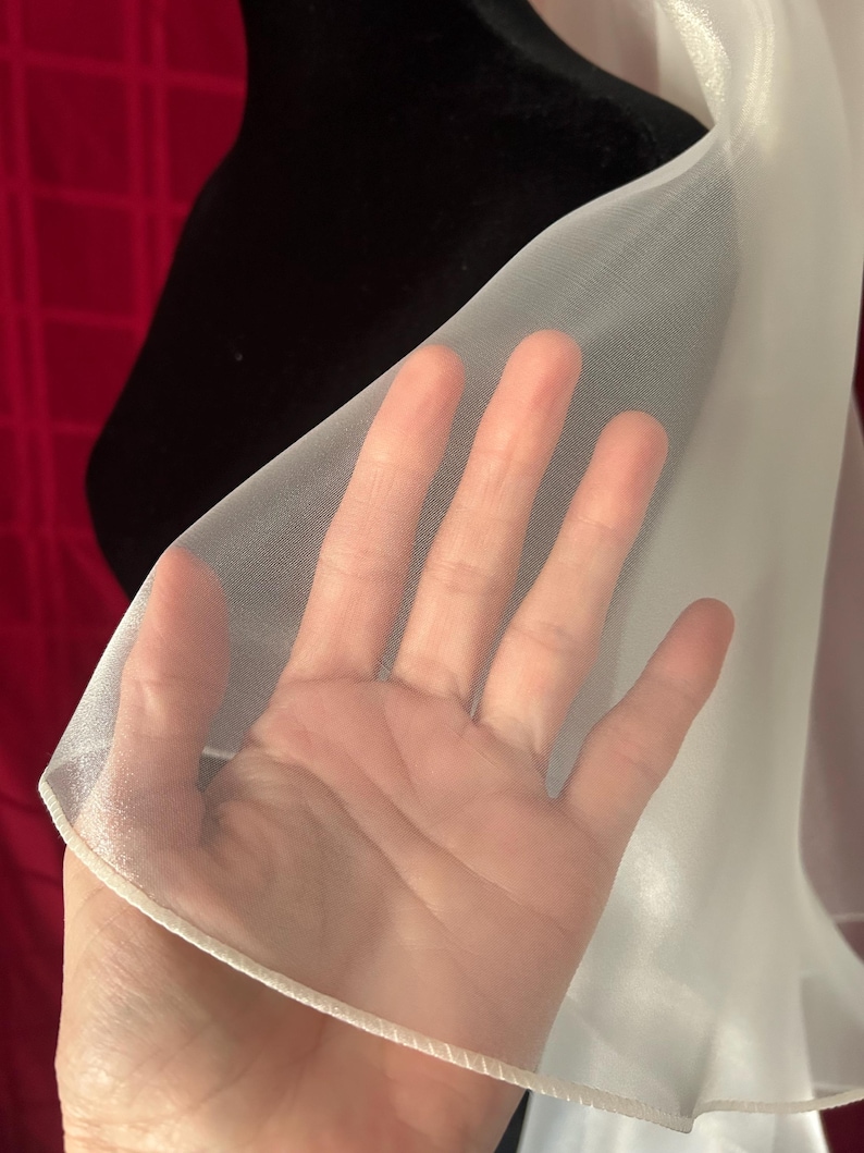 Op de afbeelding: Een close-up van een doorschijnende witte stof, mogelijk een sjaal of omslagdoek, tegen een zwarte achtergrond. De stof is dun en doorschijnend, met een hand erachter zichtbaar. De randen zijn afgewerkt met een delicate witte bies.