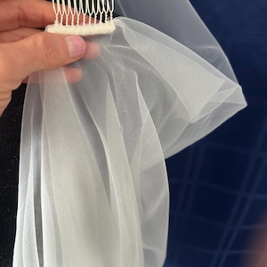 Puede incluir: Un velo de novia blanco con un peine. El velo est&aacute; hecho de una tela fina y ligera, y el peine tiene m&uacute;ltiples dientes para una sujeci&oacute;n segura en el cabello. El velo est&aacute; drapeado, mostrando su dise&ntilde;o fluido.