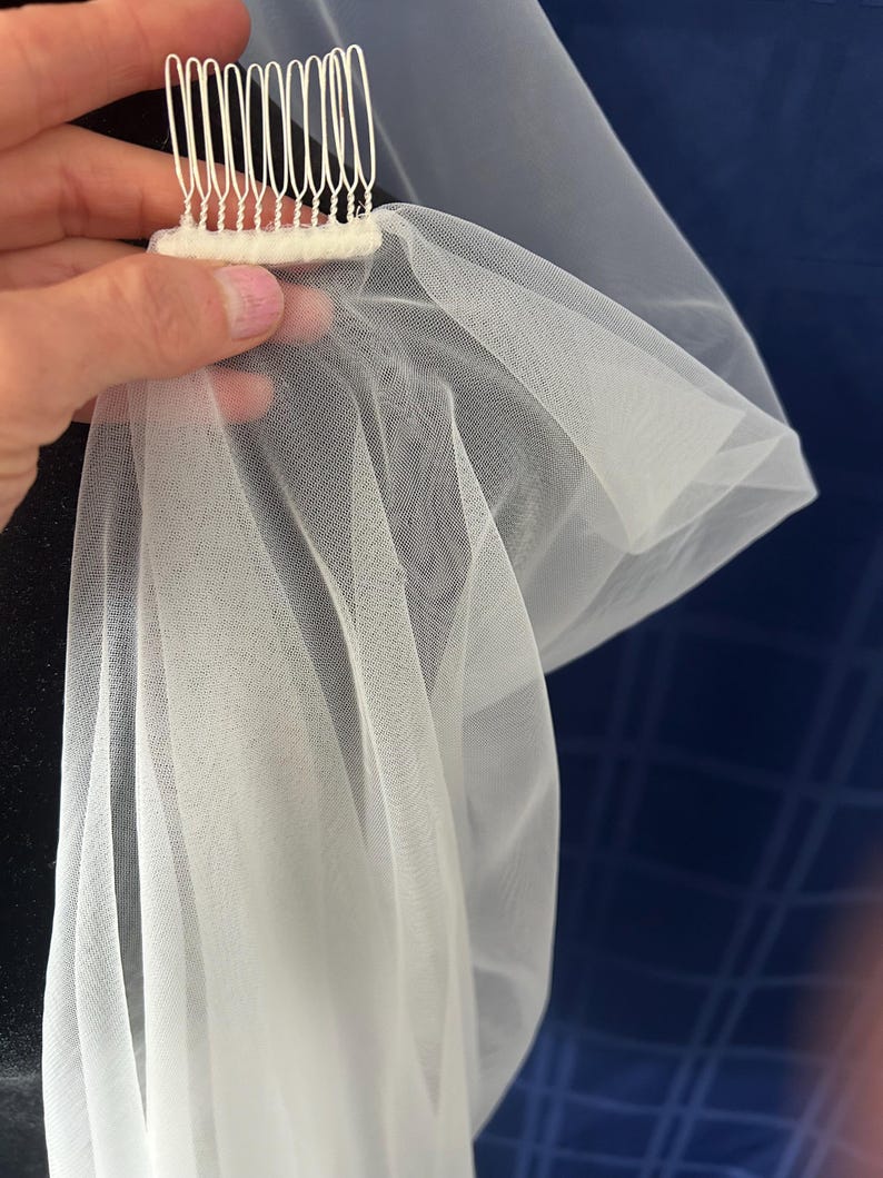 Puede incluir: Un velo de novia blanco con un peine. El velo est&aacute; hecho de una tela fina y ligera, y el peine tiene m&uacute;ltiples dientes para una sujeci&oacute;n segura en el cabello. El velo est&aacute; drapeado, mostrando su dise&ntilde;o fluido.