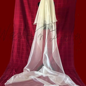 Op de afbeelding: Een gelaagde, ivoorkleurige bruidssluier. De sluier is gedrapeerd over een zwarte vorm, met een lang, vloeiend ontwerp. De stof lijkt doorschijnend en lichtgewicht, met een subtiele glans. De achtergrond is een dieprode stof.