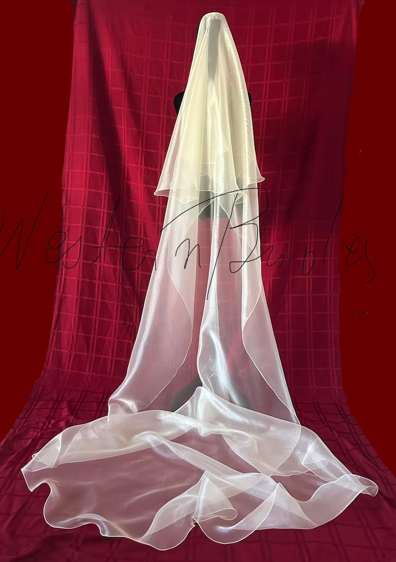 Op de afbeelding: Een gelaagde, ivoorkleurige bruidssluier. De sluier is gedrapeerd over een zwarte vorm, met een lang, vloeiend ontwerp. De stof lijkt doorschijnend en lichtgewicht, met een subtiele glans. De achtergrond is een dieprode stof.