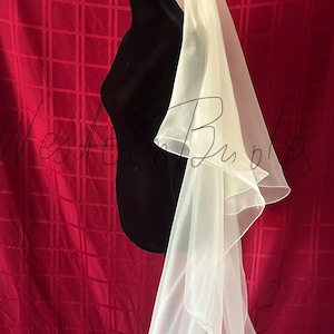 Op de afbeelding: Een witte bruidssluier met een gelaagd ontwerp, gedrapeerd over een zwarte mannequin. De sluier is gemaakt van een transparante, vloeiende stof. De achtergrond is een rode stof met een rasterpatroon.
