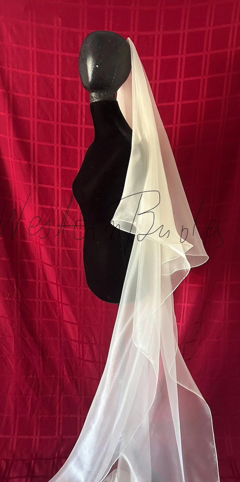 Op de afbeelding: Een witte bruidssluier met een gelaagd ontwerp, gedrapeerd over een zwarte mannequin. De sluier is gemaakt van een transparante, vloeiende stof. De achtergrond is een rode stof met een rasterpatroon.