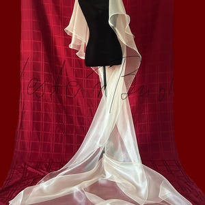 Op de afbeelding: Een ivoorkleurige bruidssluier, gedrapeerd over een zwarte mannequin. De sluier heeft een gelaagd ontwerp met een lange sleep, tegen een rode achtergrond. De stof lijkt transparant en licht.