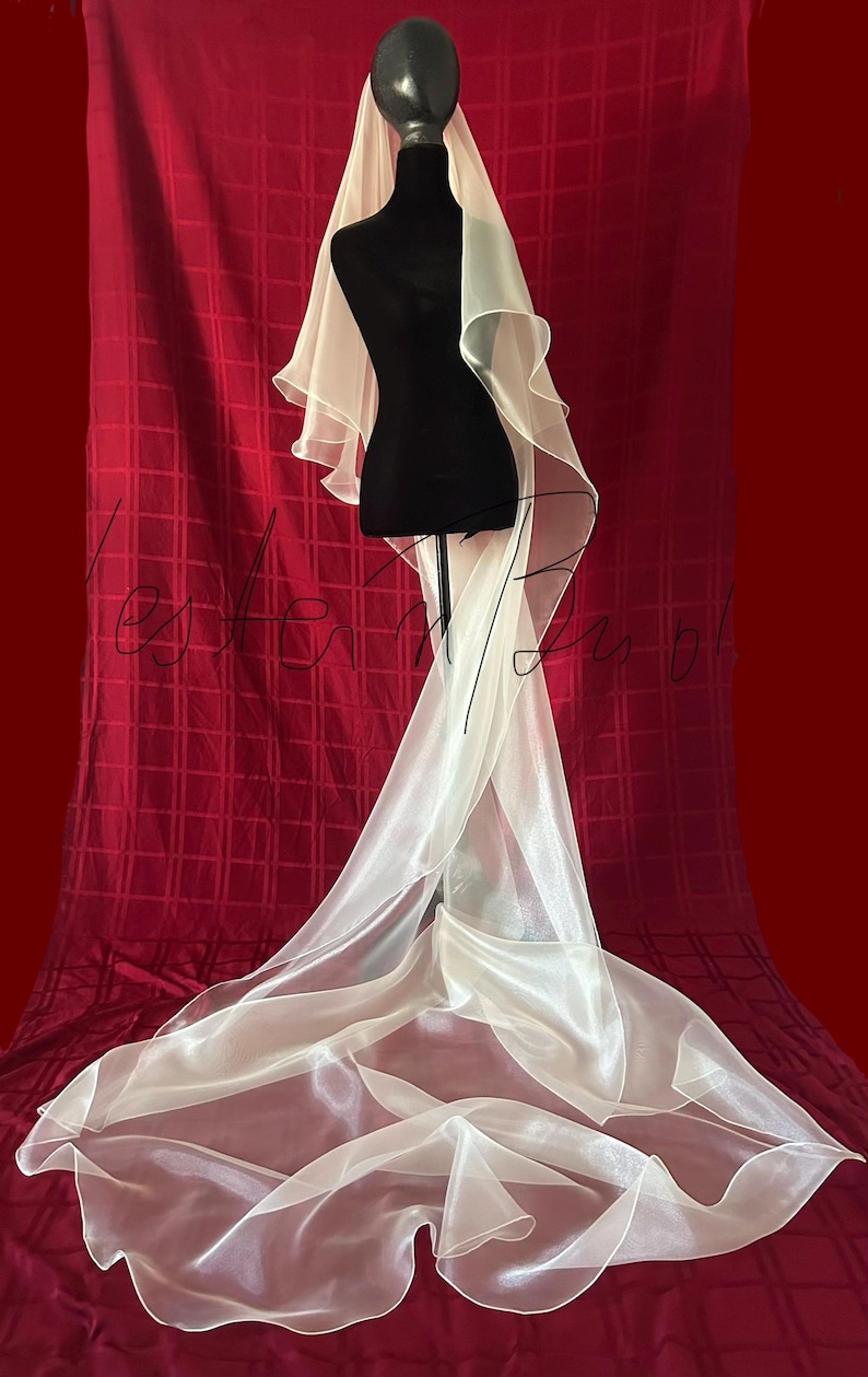 Op de afbeelding: Een ivoorkleurige bruidssluier, gedrapeerd over een zwarte mannequin. De sluier heeft een gelaagd ontwerp met een lange sleep, tegen een rode achtergrond. De stof lijkt transparant en licht.