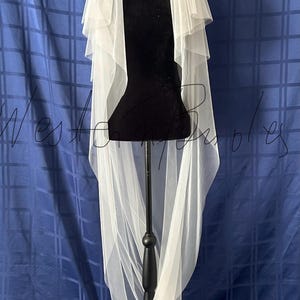 Puede incluir: Un velo de novia blanco con m&uacute;ltiples capas se muestra en un maniqu&iacute; negro. El velo es transparente y fluido, con una textura delicada. El fondo es una tela azul con un patr&oacute;n de cuadr&iacute;cula.