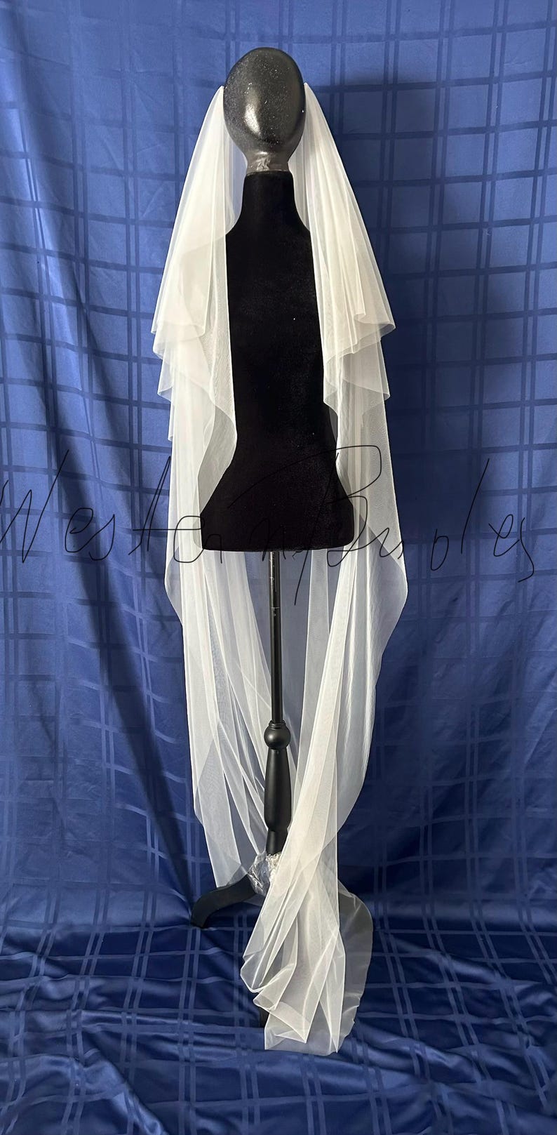 Puede incluir: Un velo de novia blanco con m&uacute;ltiples capas se muestra en un maniqu&iacute; negro. El velo es transparente y fluido, con una textura delicada. El fondo es una tela azul con un patr&oacute;n de cuadr&iacute;cula.