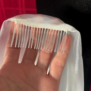 Op de afbeelding: Een close-up van een witte bruidssluier met een kam eraan. De sluier is gemaakt van een transparante, vloeiende stof. De kam heeft verschillende doorzichtige plastic tanden. De achtergrond is donker.