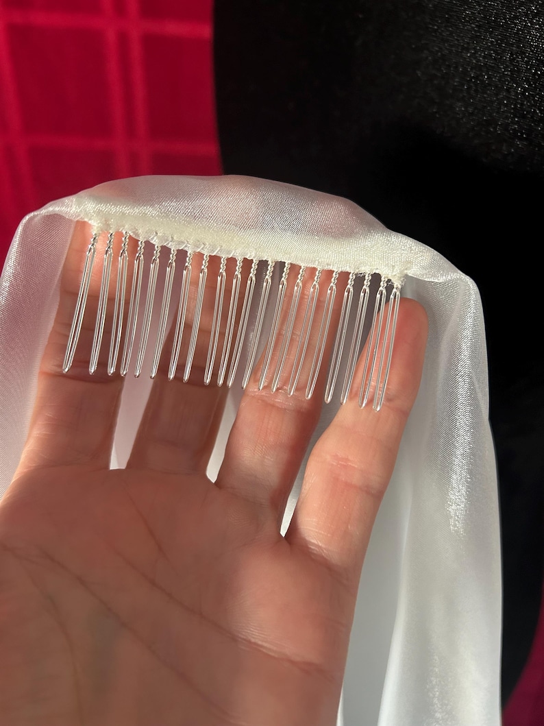 Op de afbeelding: Een close-up van een witte bruidssluier met een kam eraan. De sluier is gemaakt van een transparante, vloeiende stof. De kam heeft verschillende doorzichtige plastic tanden. De achtergrond is donker.