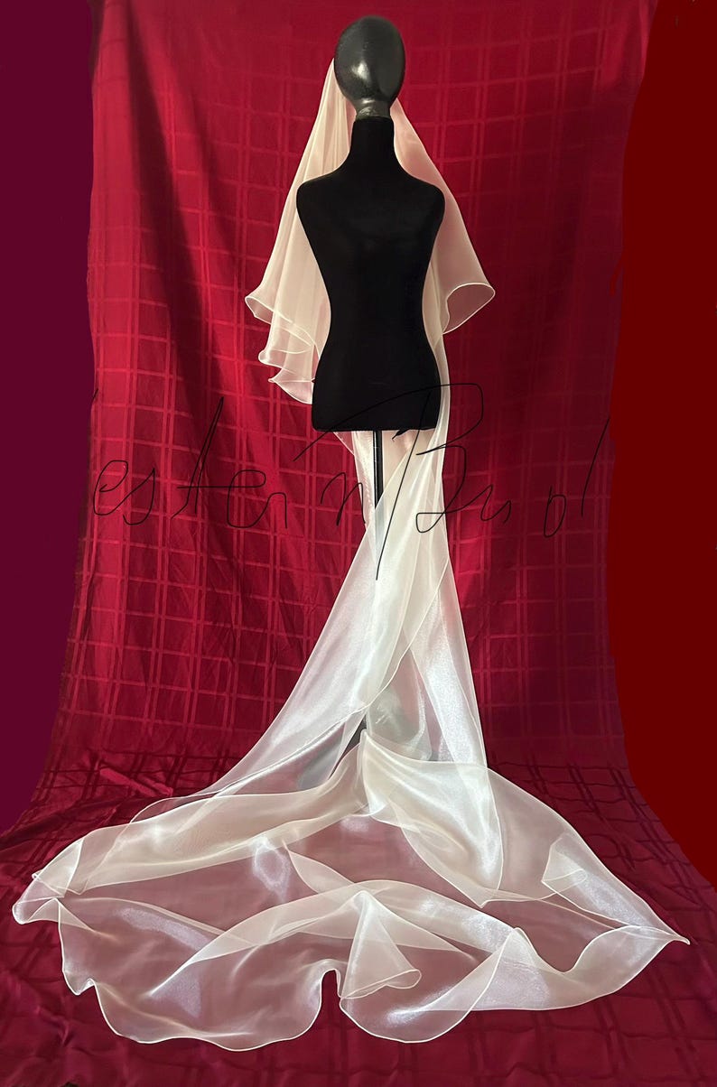 Op de afbeelding: Een ivoorkleurige bruidssluier is gedrapeerd over een zwarte mannequin. De sluier heeft een lange sleep en gelaagde details bij de schouders. De achtergrond is een dieprode stof met een rasterpatroon.