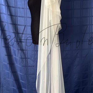 Puede incluir: Un velo de novia blanco con m&uacute;ltiples capas de tela suave y transparente. El velo est&aacute; drapeado sobre una cabeza y hombros de maniqu&iacute; negro, mostrando su dise&ntilde;o fluido. El fondo es una tela azul.